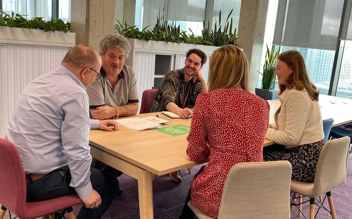 Photo in a brightly lit office. Five people are sat around a desk smiling and in conversation.